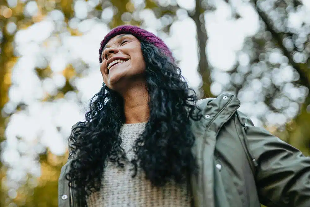 Woman smiling outdoors with trees behind her, natural light highlighting warmth and ease