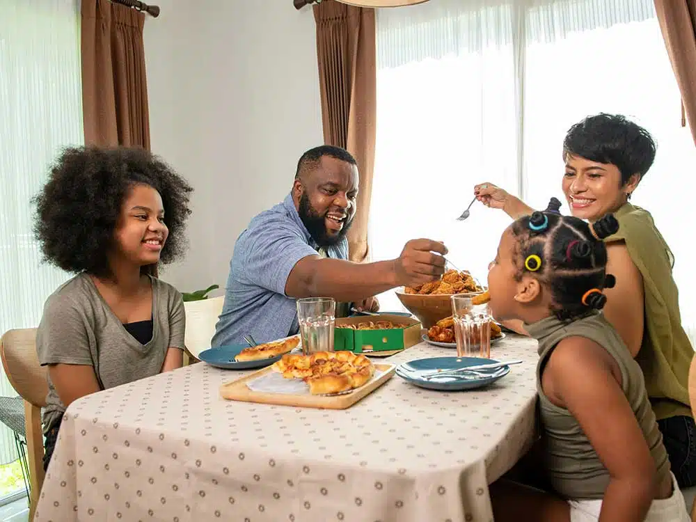 An African American father is feeding his daughters dinner at the table while the whole family laughs together during a fun evening.