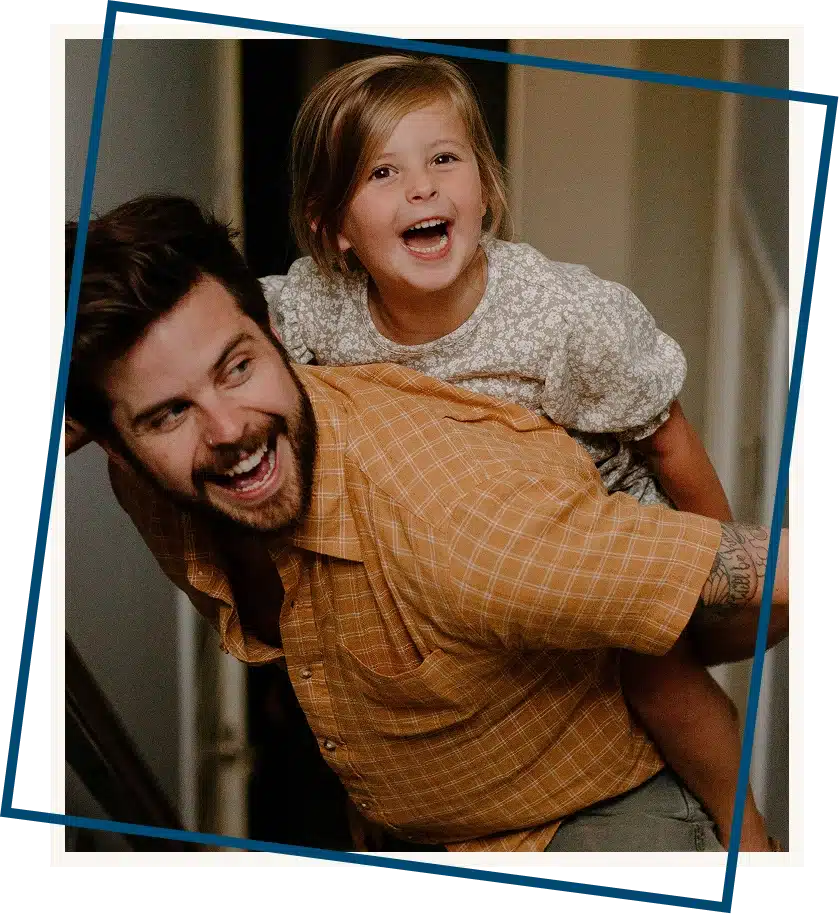 Father laughing while giving his child a piggyback ride, both smiling during a playful family moment outside in the sunshine