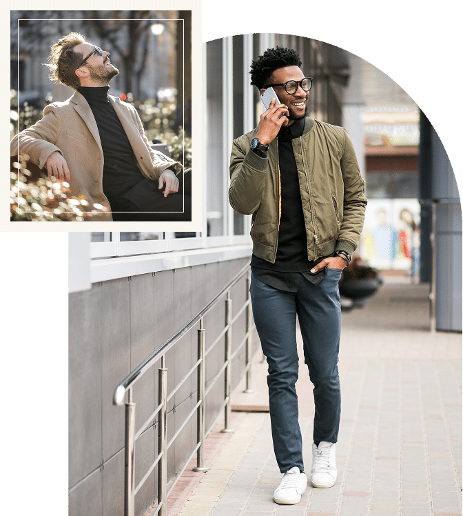 A smiling man sitting on a bench looking up while another man walks and talks on the phone