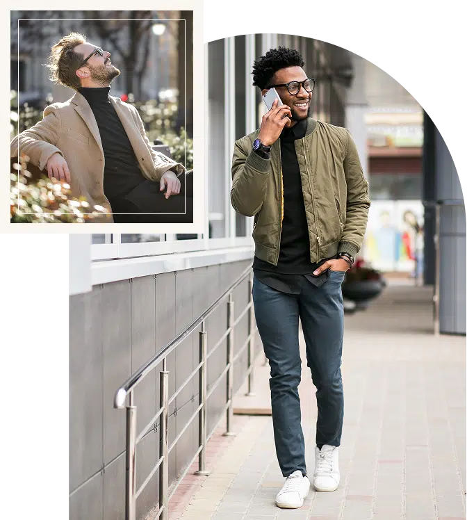A smiling man sitting on a bench looking up while another man walks and talks on the phone
