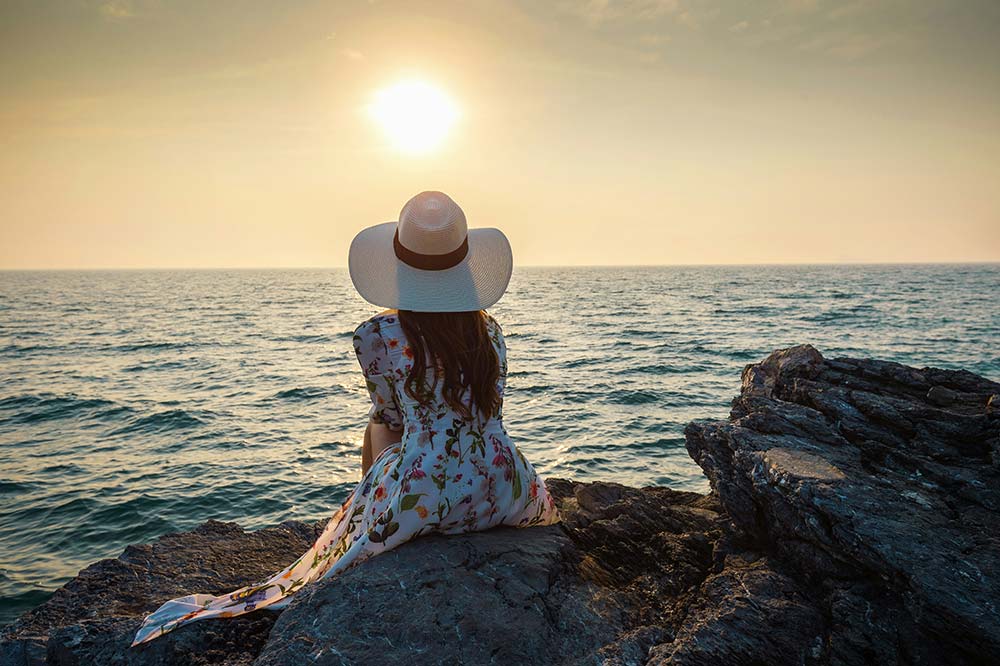 A woman in a floral dress and a wide-brimmed sun hat sits on a rocky cliff, looking out at the ocean as the sun sets over the water.