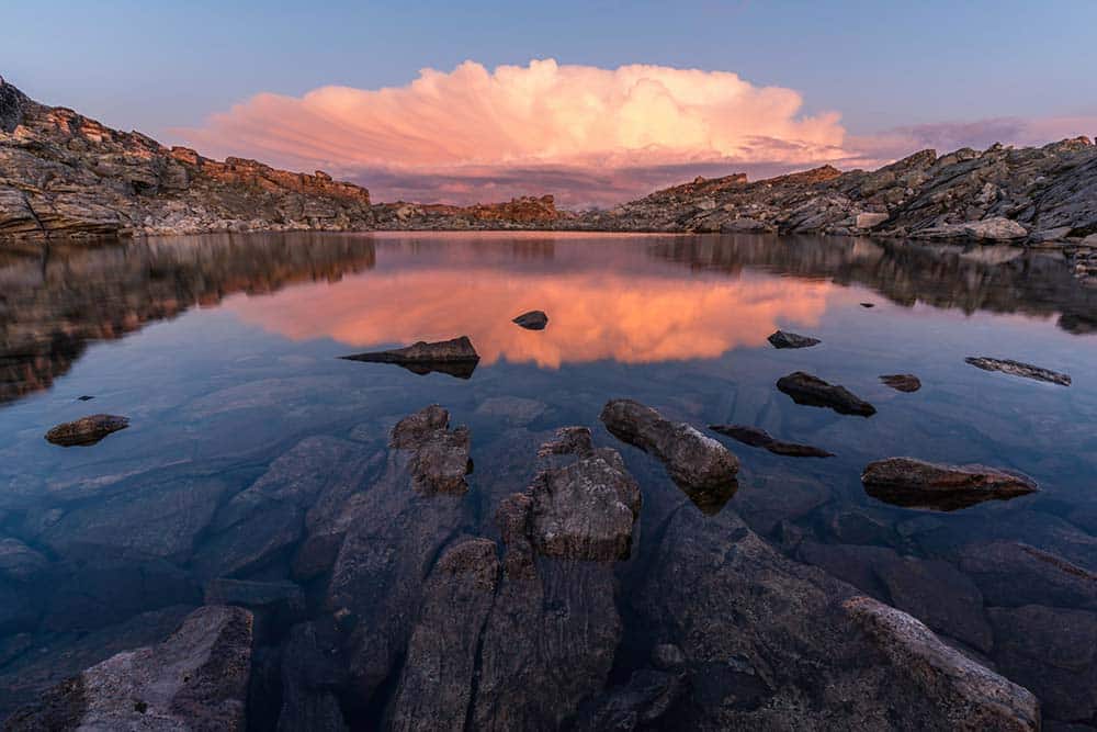 A still, clear mountain lake reflects a massive, glowing pink cloud at sunset.
