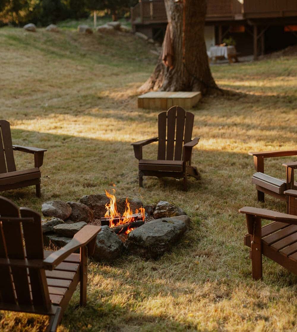 A campfire burning in a forest clearing with empty chairs arranged around it