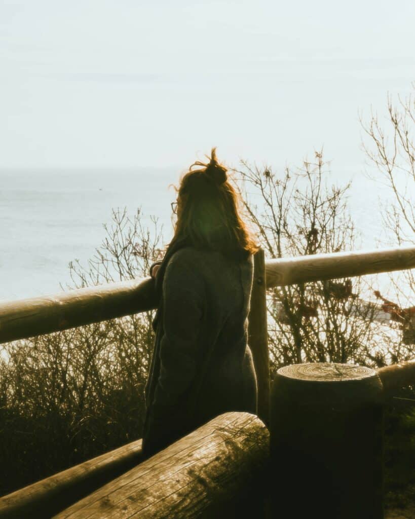 A woman standing quietly by a wooden overlook, facing a sunlit body of water