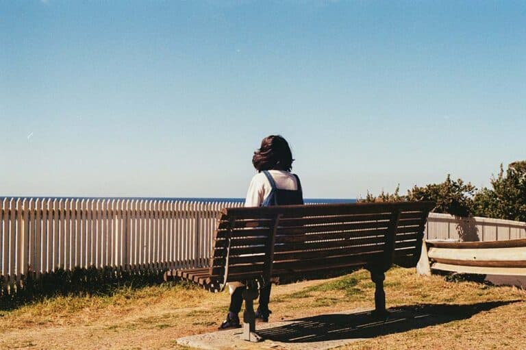 A woman sitting on a bench by the ocean, looking out at the water and horizon in a peaceful coastal setting.