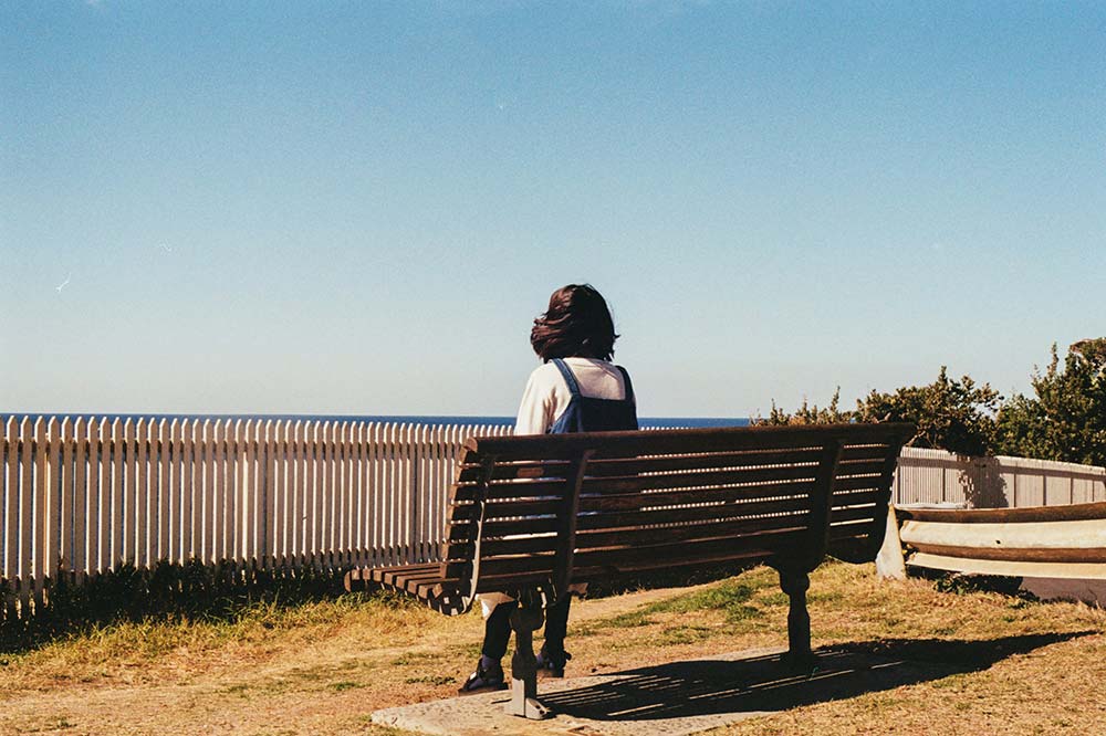 A woman sitting on a bench by the ocean, looking out at the water and horizon in a peaceful coastal setting.